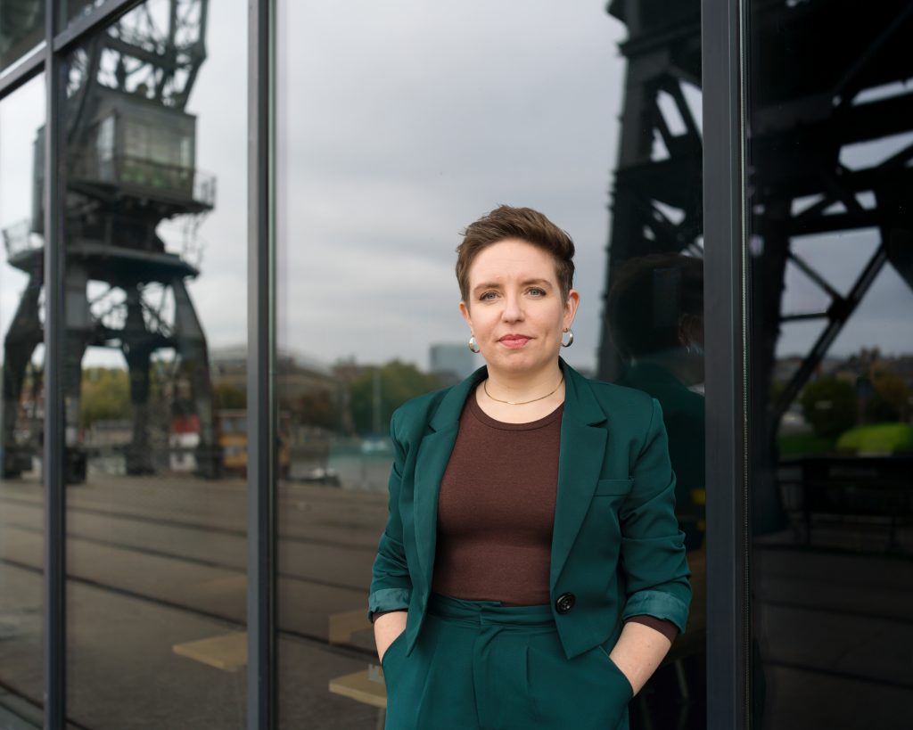 Carla Denyer stands looking at the camera against a large glass wall.